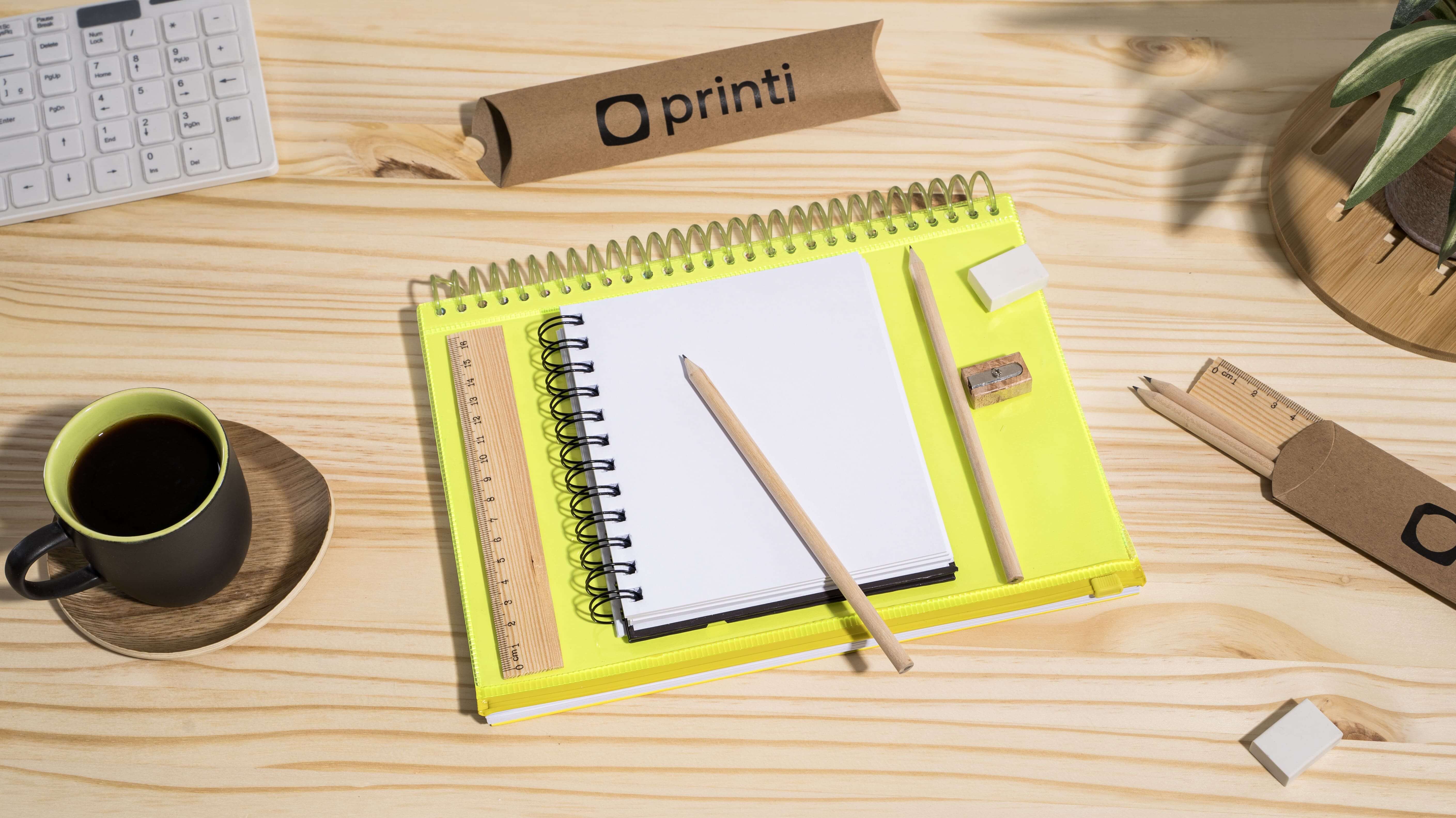 Imagem vista de cima mostrando papelaria corporativa composta por caderno, lapis, regua, borracha, um kit de escrita completo. Os objetos estão ao lado de uma xícara de café e em cima de uma mesa de madeira e