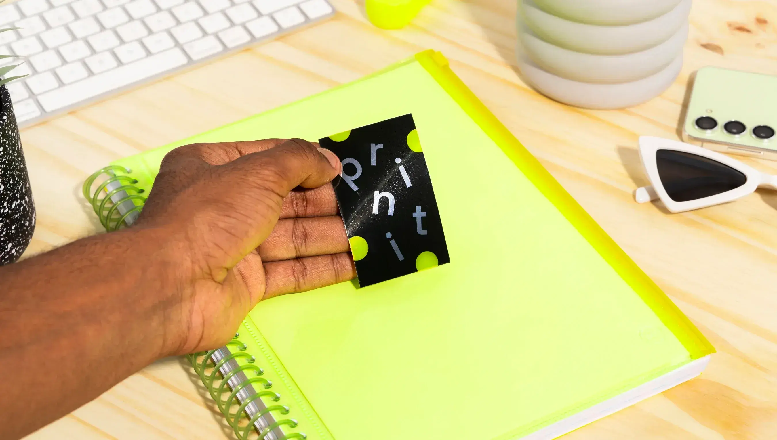 Mão de uma pessoa negra segurando um adesivo retangular perosnalizado com o nome e cores da Printi, em  cima de um caderno verde apoiado em uma superfície de madeira