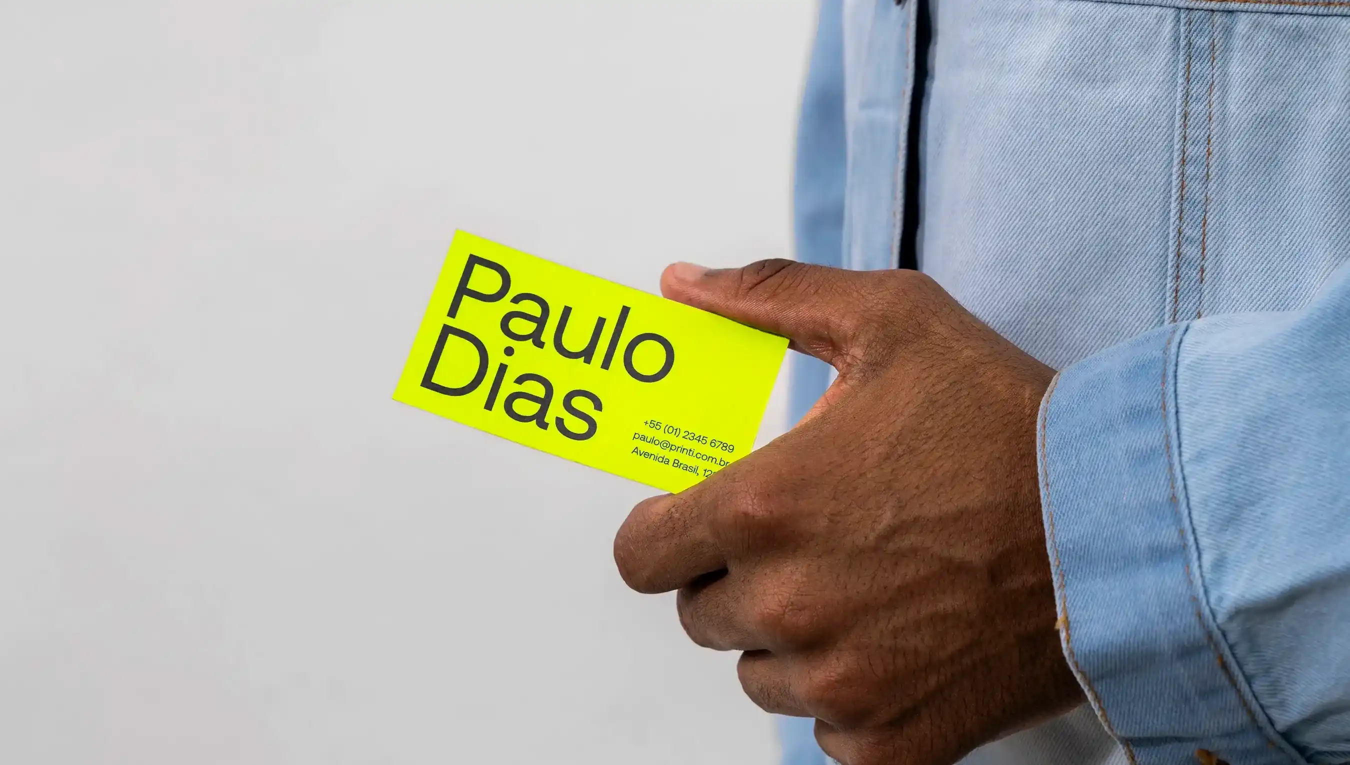 Destaque na mão de um homem negro usando uma jaqueta jeans segurando um cartão de visita verde com o nome paulo dias