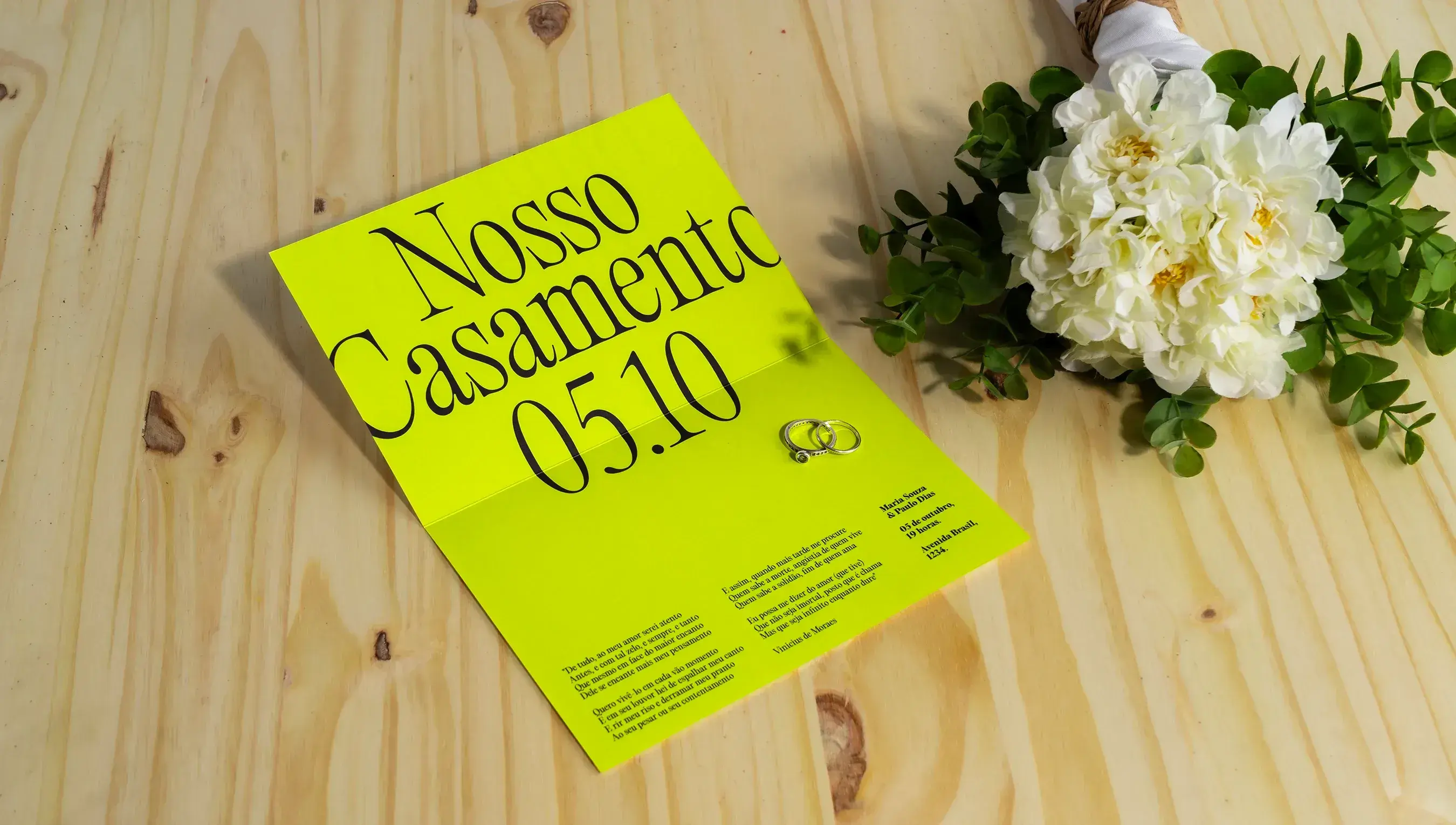 Convite de casamento na cor verde e com a fonte na cor preta. O convite está em cima de uma mesa de madeira, com uma flor ao lado
