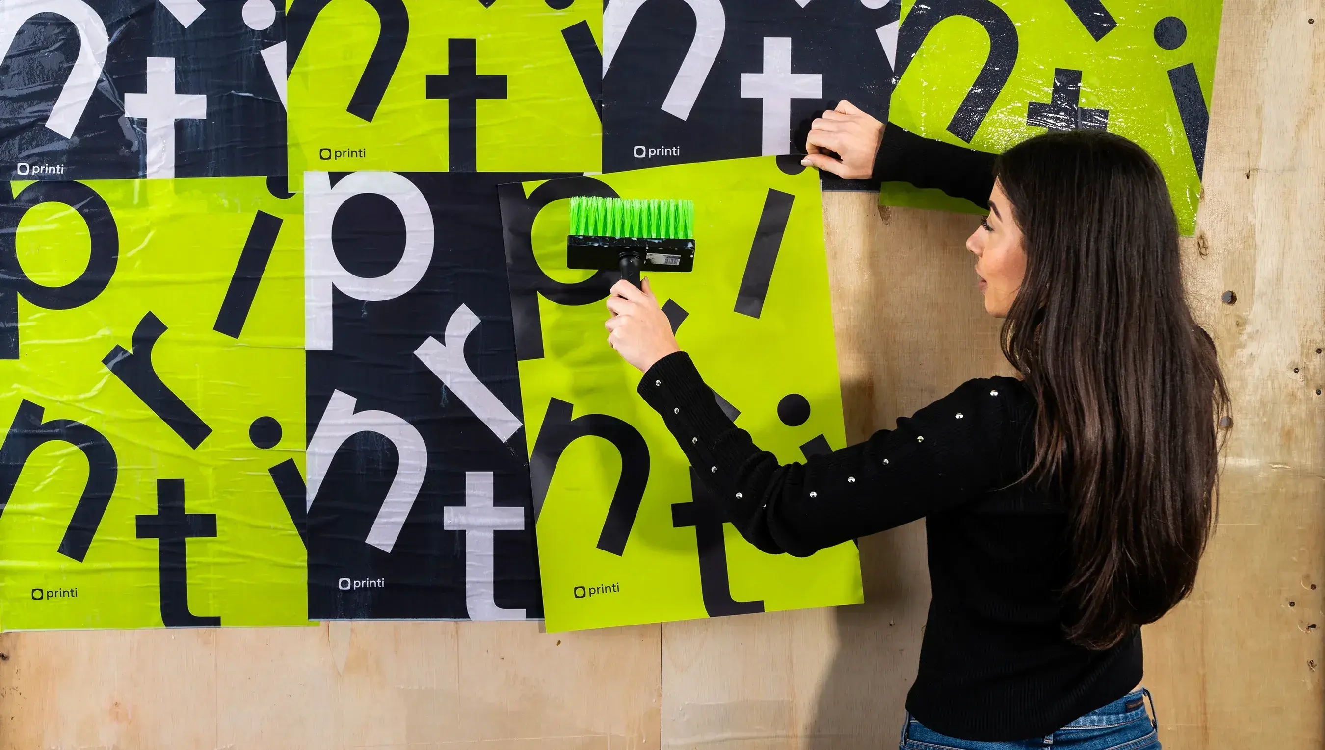 Mulher de costas usando blusa presta fazendo a aplicação do produto lambe lambe em uma parede. O lambe lambe está personalizado com o nome da printi mesclando entre as cores pretas e verde