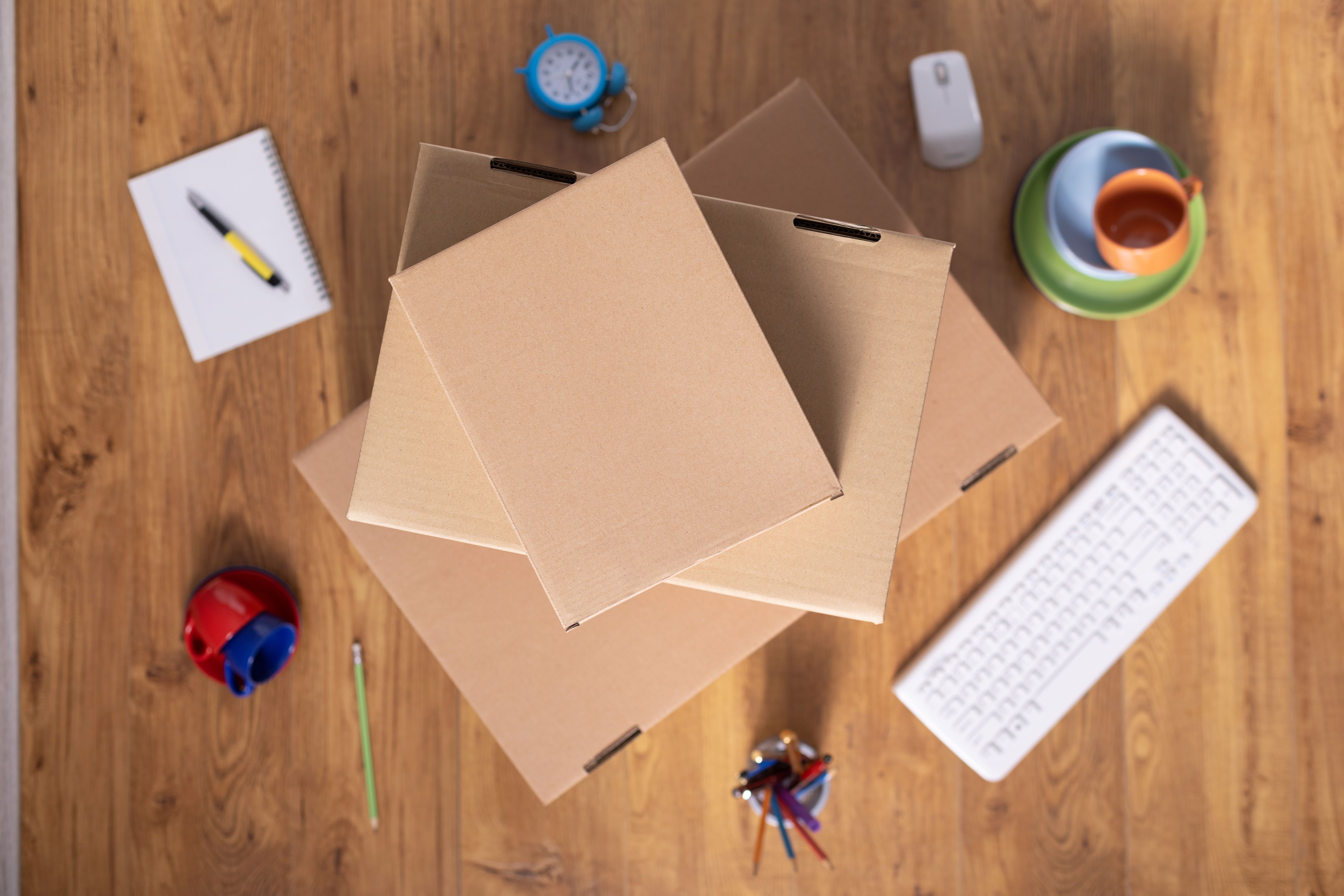 Vista superior de caixas de papelão empilhadas sobre mesa de escritório com um teclado branco, caderno, relogio e outros itens